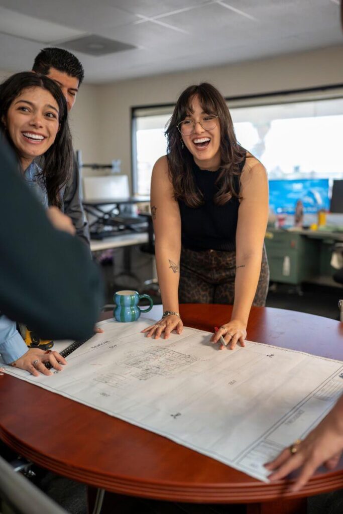 Smiling team members gathered around a table reviewing large architectural drawings.